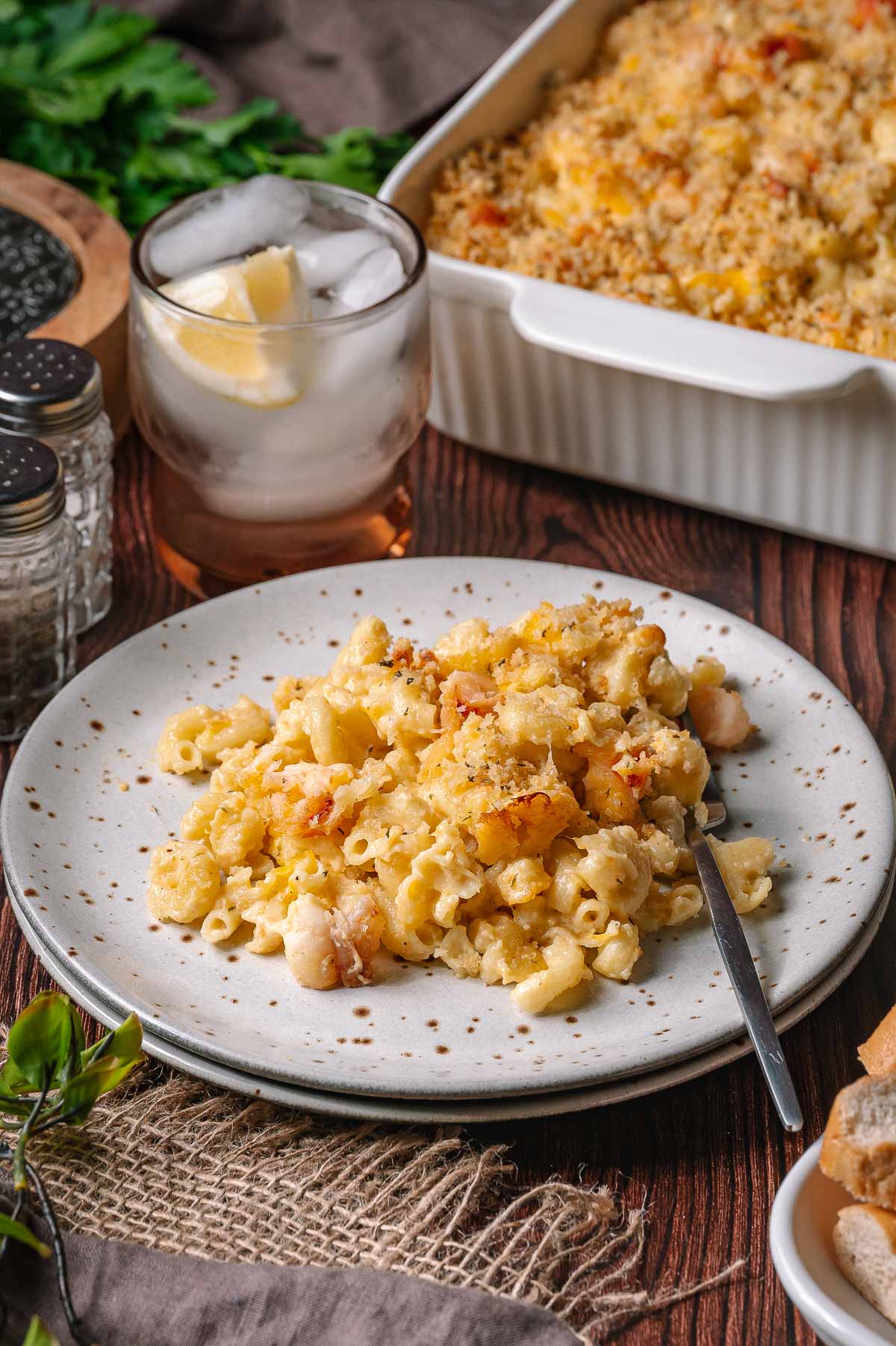 A plate of baked lobster mac and cheese with breadcrumbs sits next to a glass of iced water and a casserole dish on a wooden table.