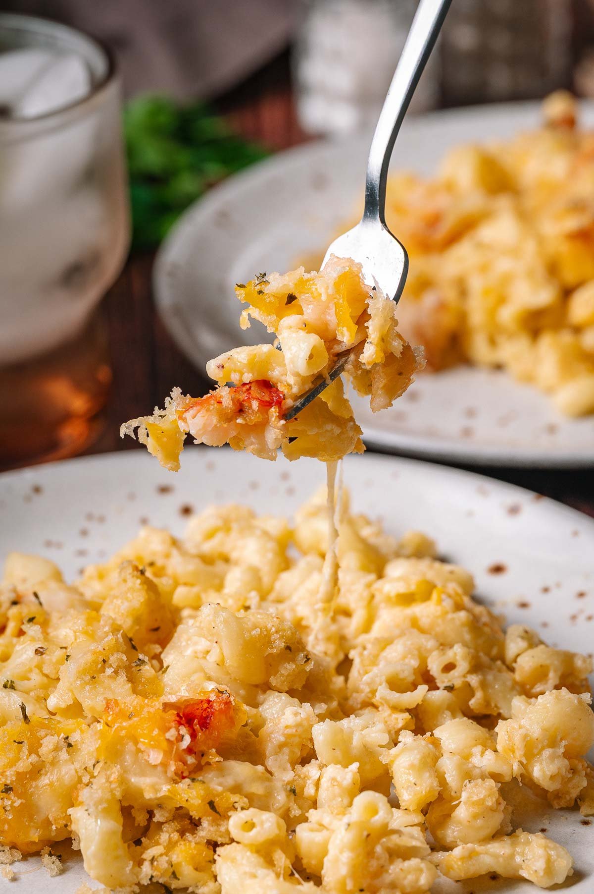 A close-up of a fork lifting a bite of baked lobster mac and cheese from a plate, with another plate and a glass of ice water in the background.