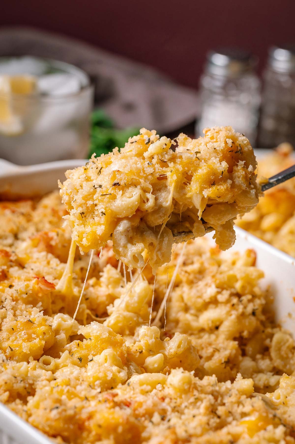 A serving of baked lobster macaroni and cheese is being lifted from a casserole dish, showing melted cheese and a crispy breadcrumb topping.