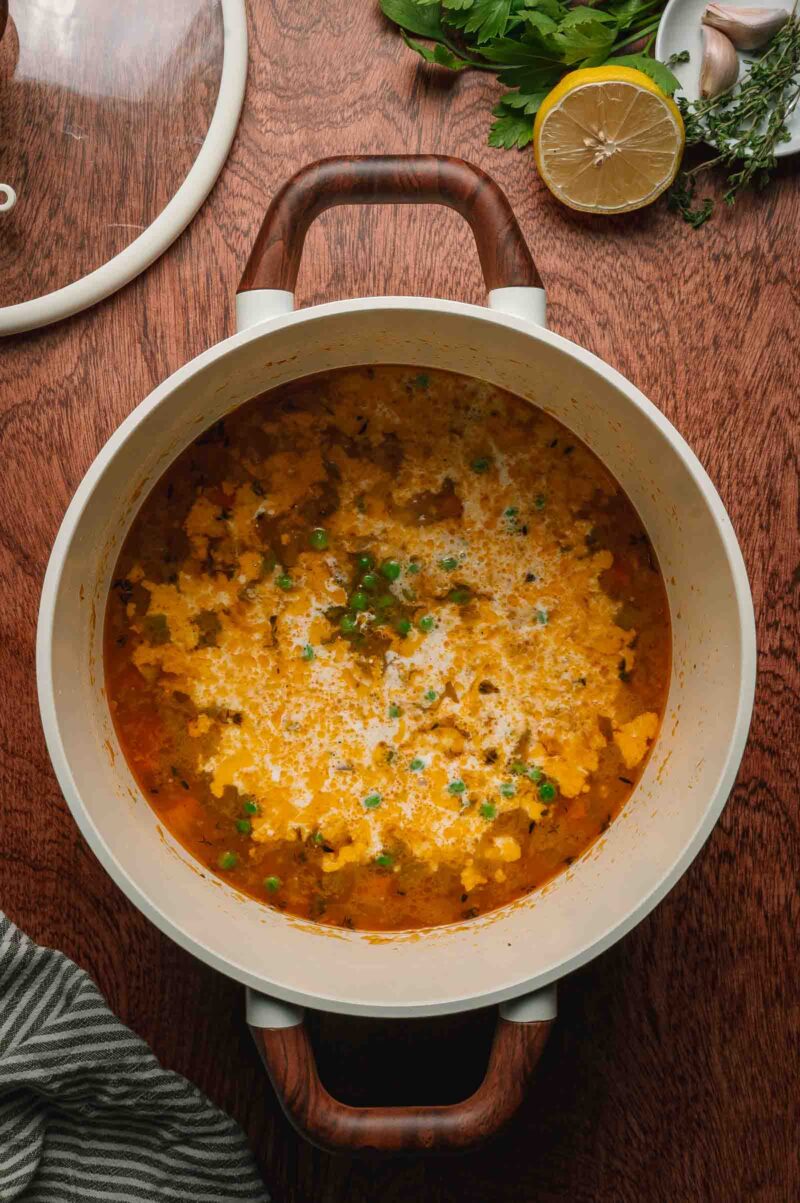A pot of pot pie base with visible peas, herbs, with cream added to the orange broth sits on a wooden table beside a lemon half, fresh herbs, and a striped cloth.