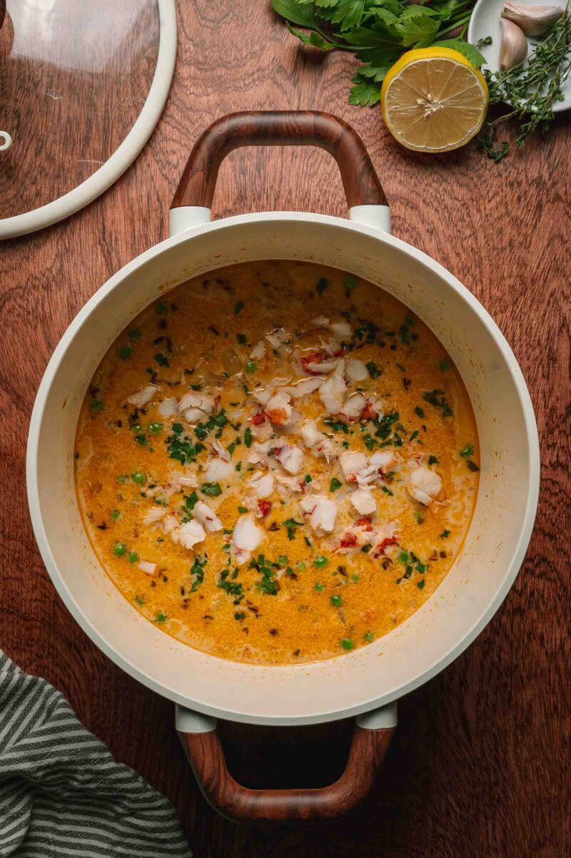 A pot of creamy pot pie filling with chopped herbs and lobster sits on a wooden table, surrounded by a lemon half, herbs, and a garlic clove.