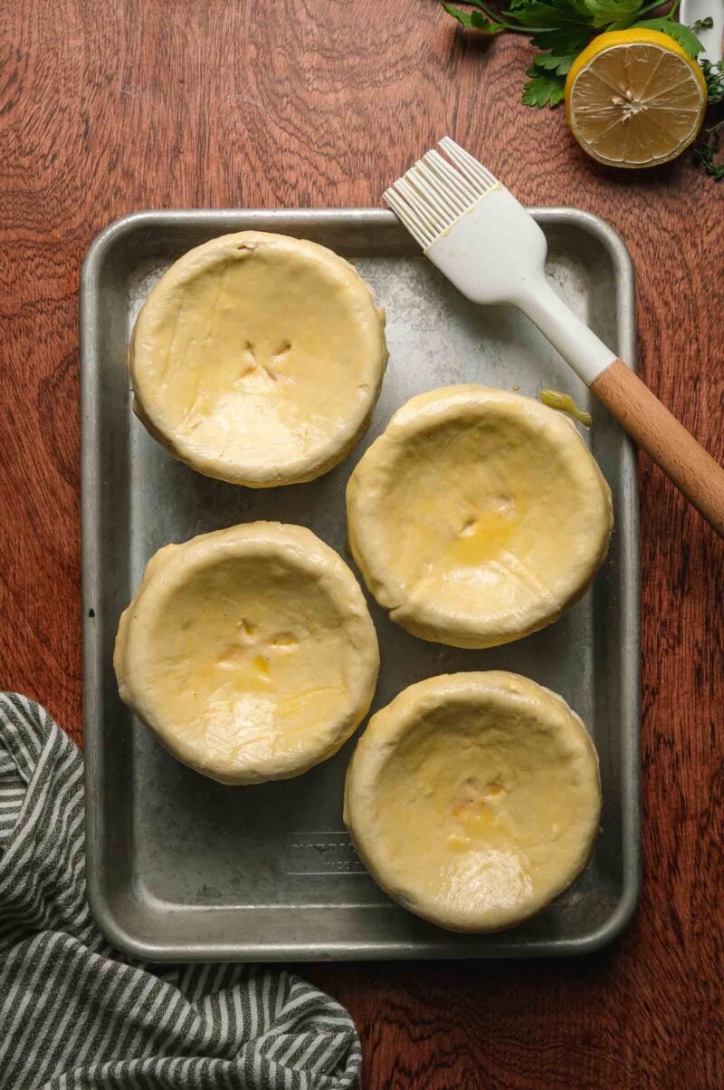 Four unbaked pot pies with golden egg wash tops sit on a metal baking tray next to a pastry brush, lemon half, parsley, and striped cloth on a wooden surface.
