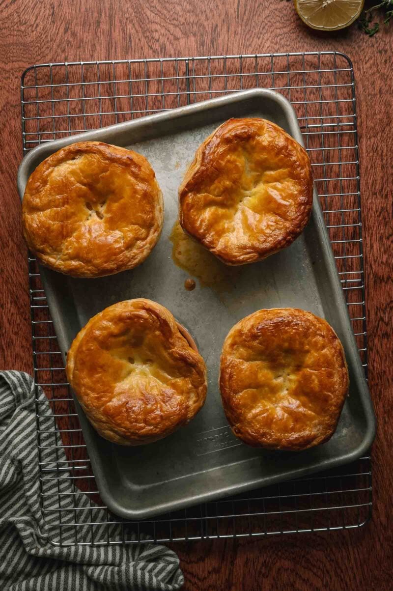 Four golden-brown lobster pot pies sit on a baking tray, which is placed on a wire rack over a wooden surface. A striped cloth and a lemon slice are visible nearby.