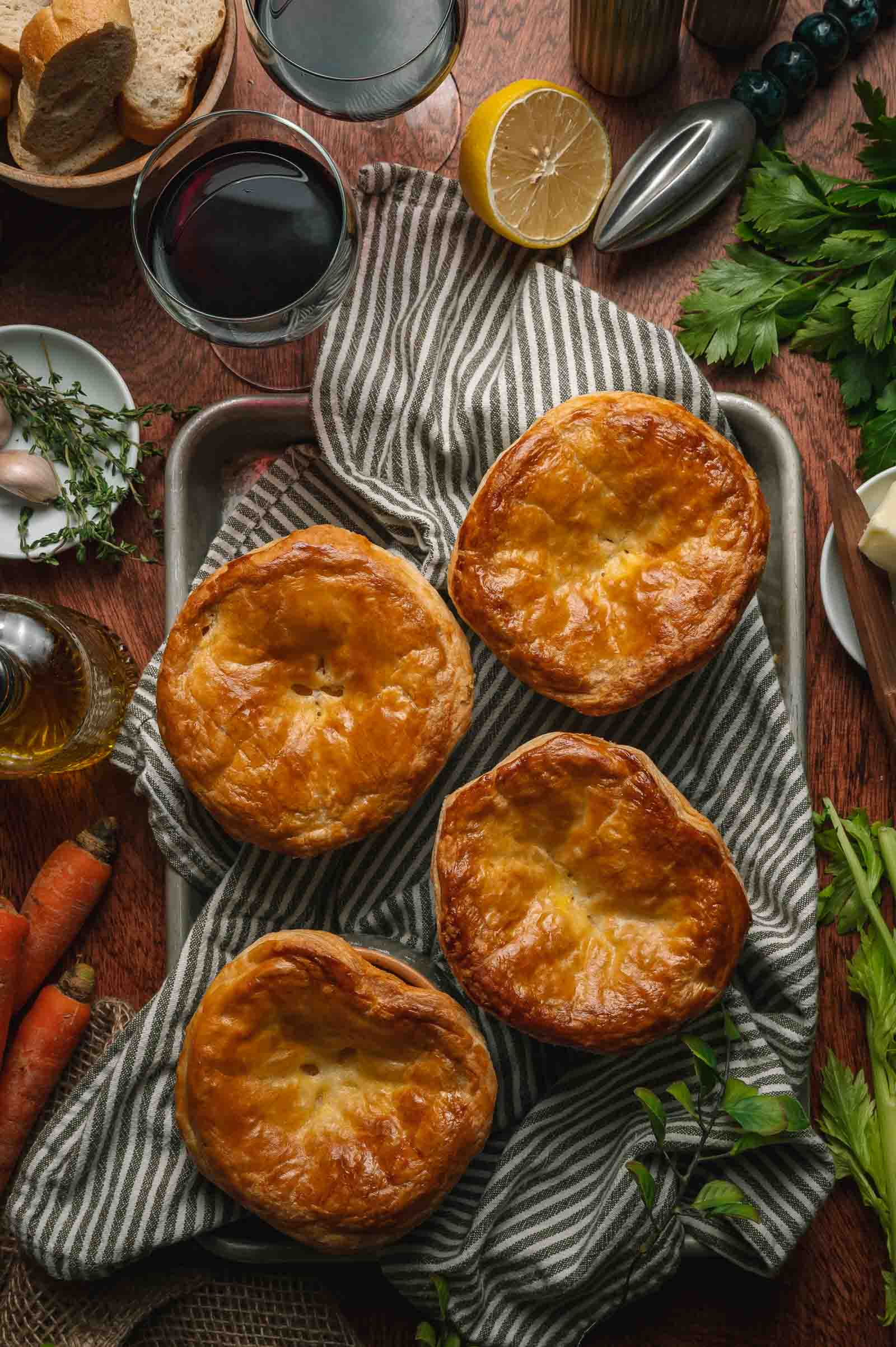 Four golden-brown lobster pot pies on a striped cloth, surrounded by fresh herbs, carrots, bread, lemon, and two glasses of red wine on a wooden table.