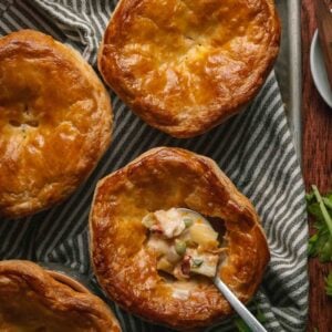 Four golden-brown lobster pot pies on a striped cloth, with one pie partially open to reveal a creamy filling and vegetables, a spoon inside the open pie.