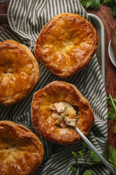 Four golden-brown lobster pot pies on a striped cloth, with one pie partially open to reveal a creamy filling and vegetables, a spoon inside the open pie.