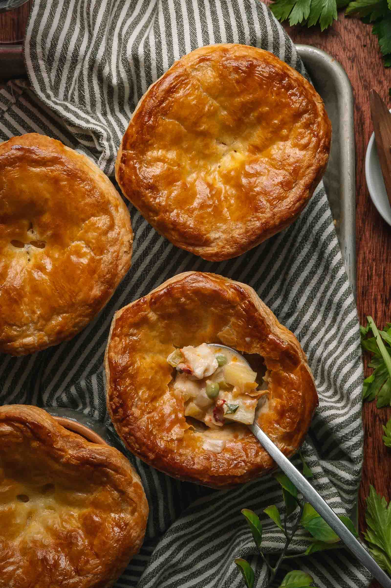 Four golden-brown lobster pot pies on a striped cloth, with one pie partially open to reveal a creamy filling and vegetables, a spoon inside the open pie.