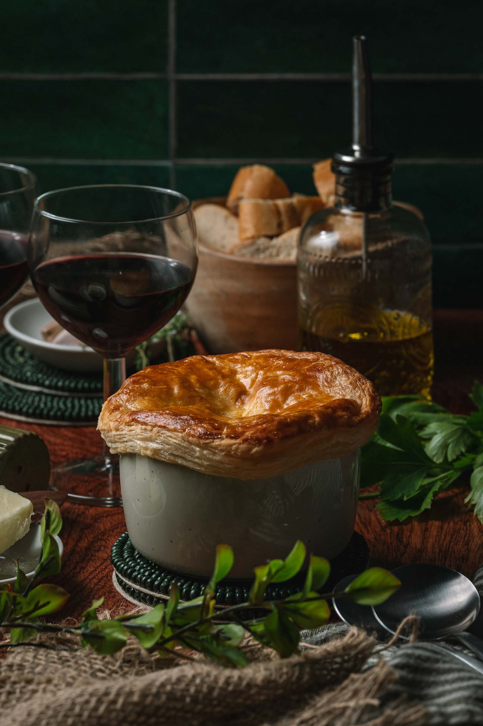 A golden-brown puff pastry lobster pot pie in a white ramekin sits on a table next to a glass of red wine, bread, olive oil, and fresh herbs.