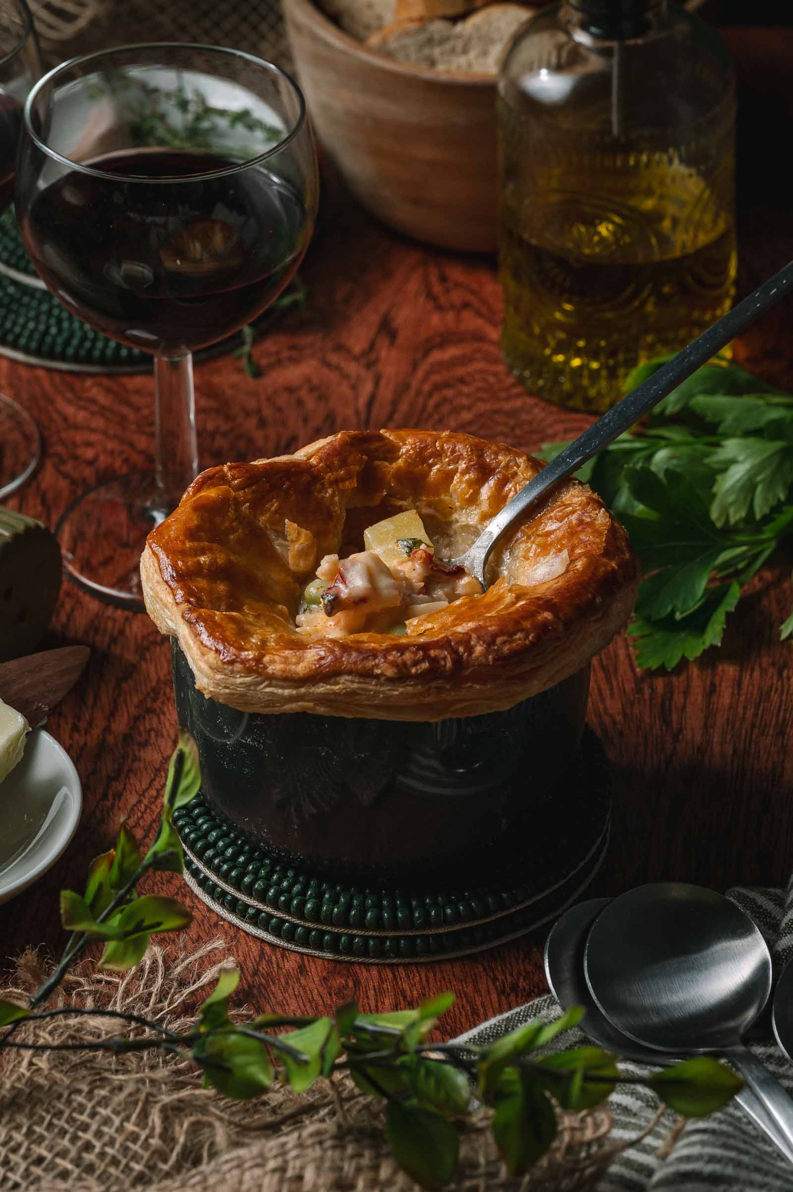A spoon rests in a baked lobster pot pie with a golden crust, set on a wooden table beside a glass of red wine, olive oil, parsley, bread, and cutlery.