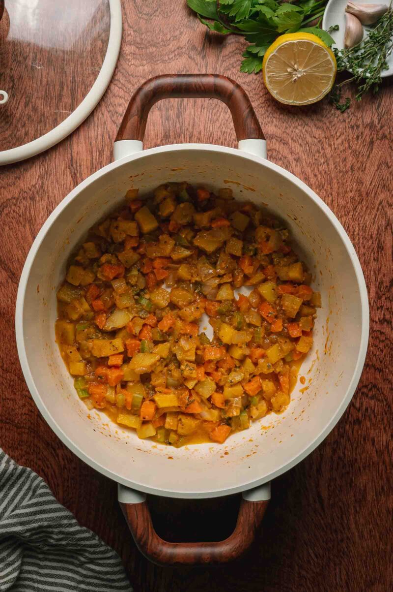 A white pot containing sautéed diced vegetables coated in cooked flour, including carrots, celery, and onions, sits on a wooden surface beside herbs, a lemon half, garlic, and a striped cloth.