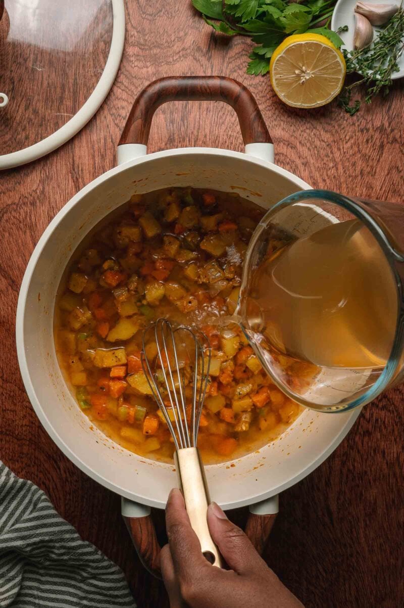 A hand pours broth from a measuring cup into a pot of diced vegetables being whisked, with herbs, garlic, and half a lemon on the table nearby.