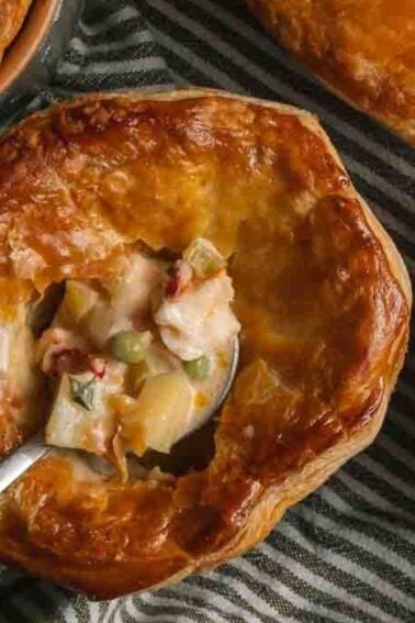 Golden-brown pot pies on a striped cloth, with one pie partially opened to reveal a creamy vegetable and meat filling being scooped with a spoon.