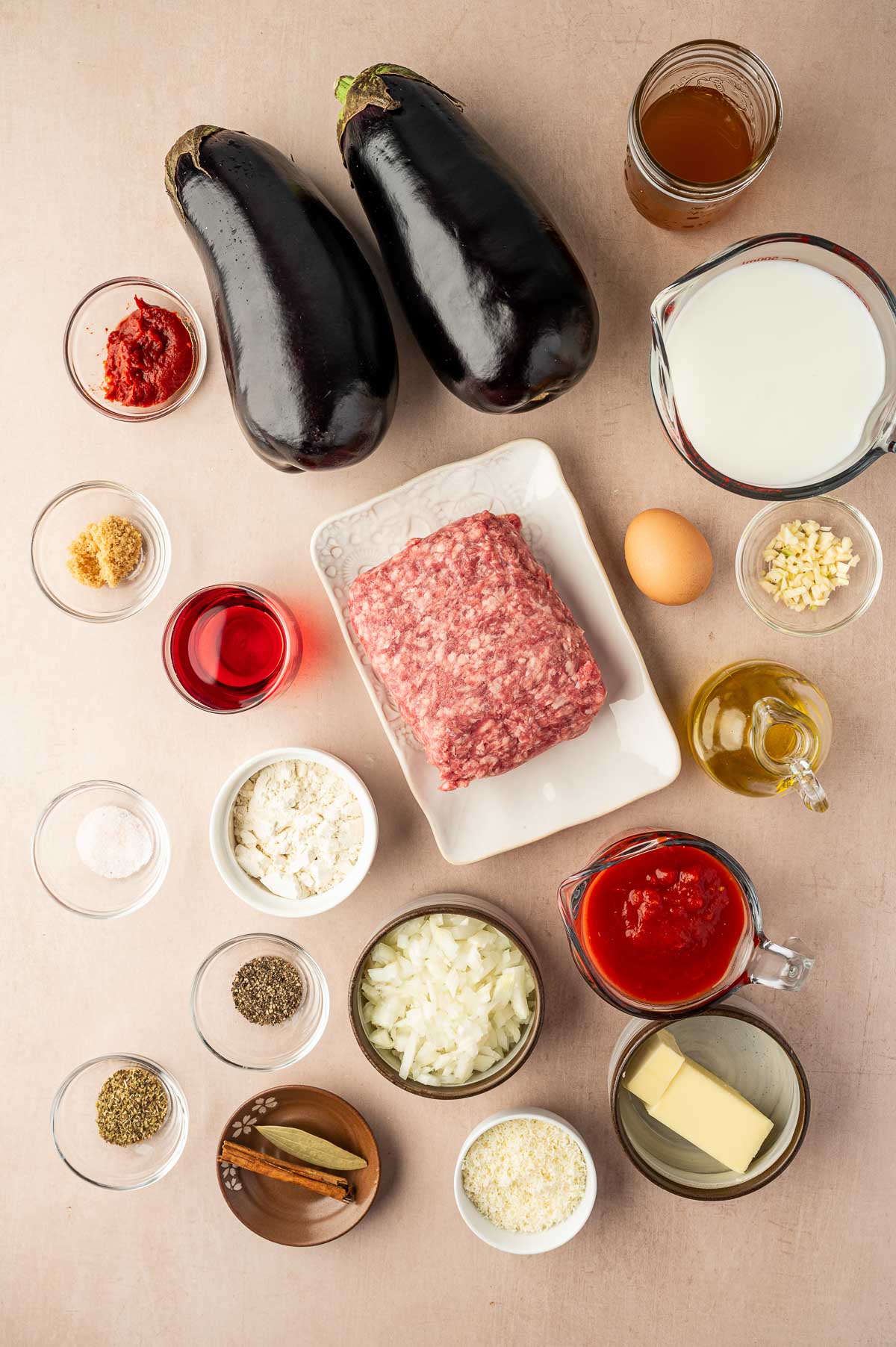 Ingredients for moussaka arranged on a table, including eggplants, ground lamb, milk, broth, tomato sauce, onion, egg, butter, spices, flour, olive oil, and grated cheese in bowls.