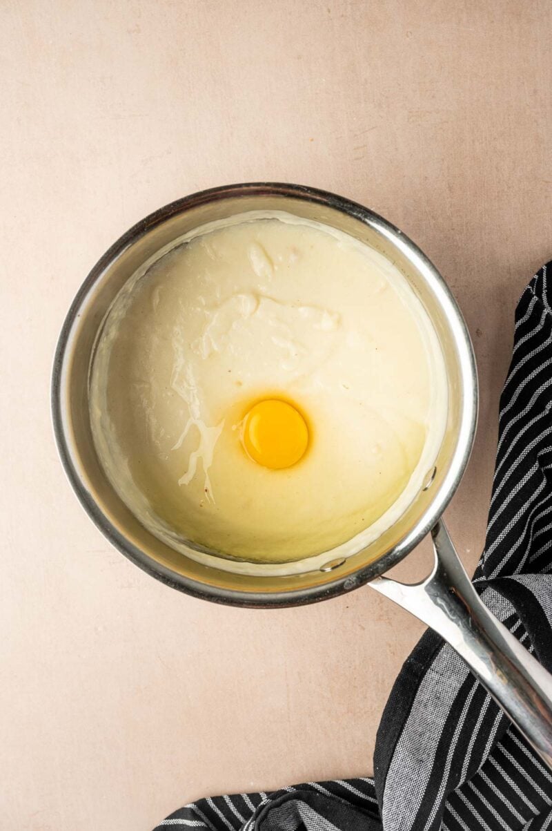 A stainless steel saucepan containing a light-colored creamy béchamel with a raw egg yolk in the center, next to a black and white striped cloth.