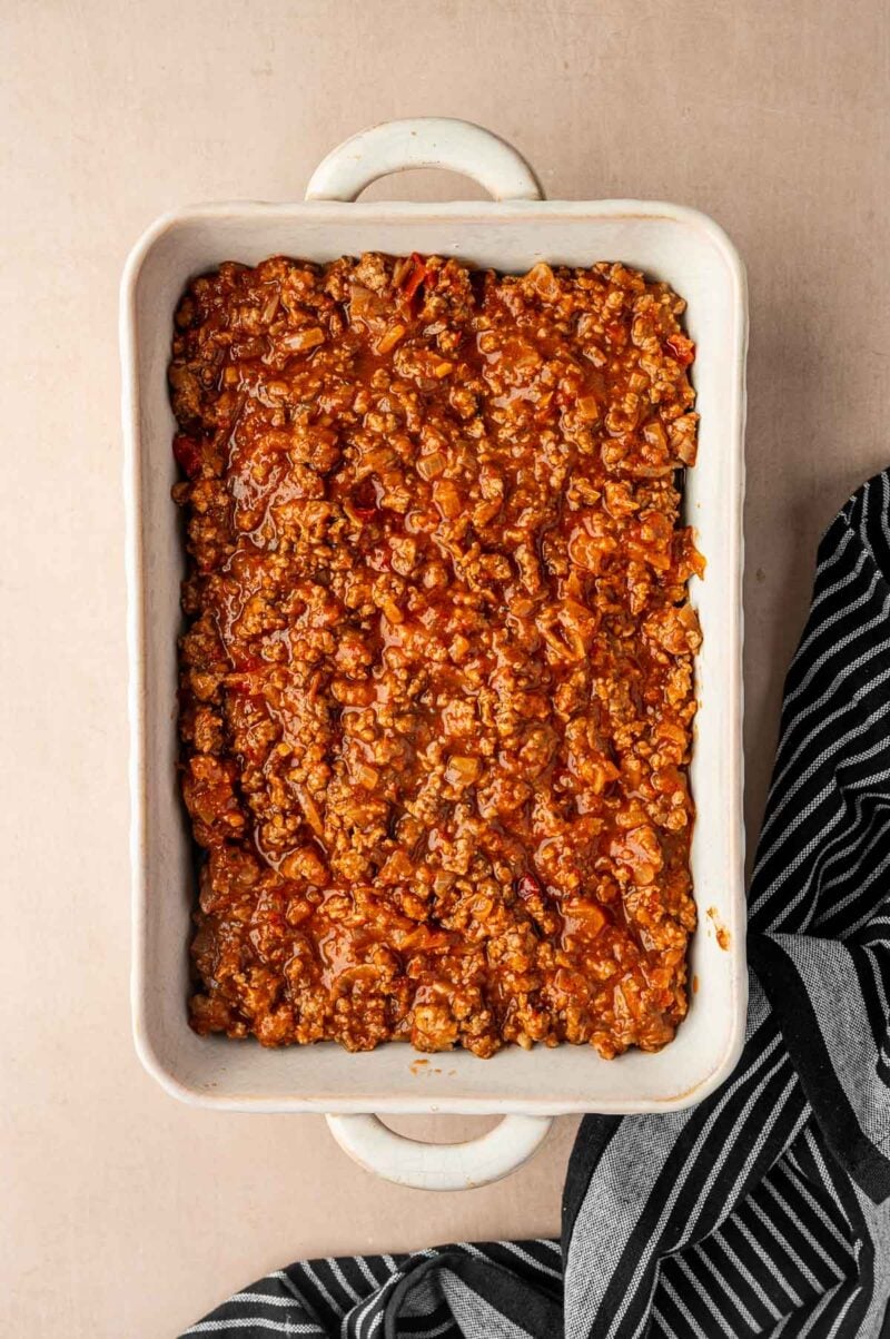 A white rectangular baking dish filled with cooked ground lamb in tomato sauce sits on a beige surface next to a black and white striped cloth.