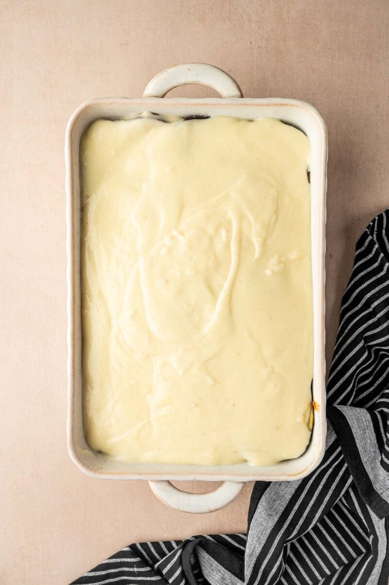 A rectangular baking dish filled with an even layer of creamy white sauce, with a striped black and white cloth partially visible beside it.