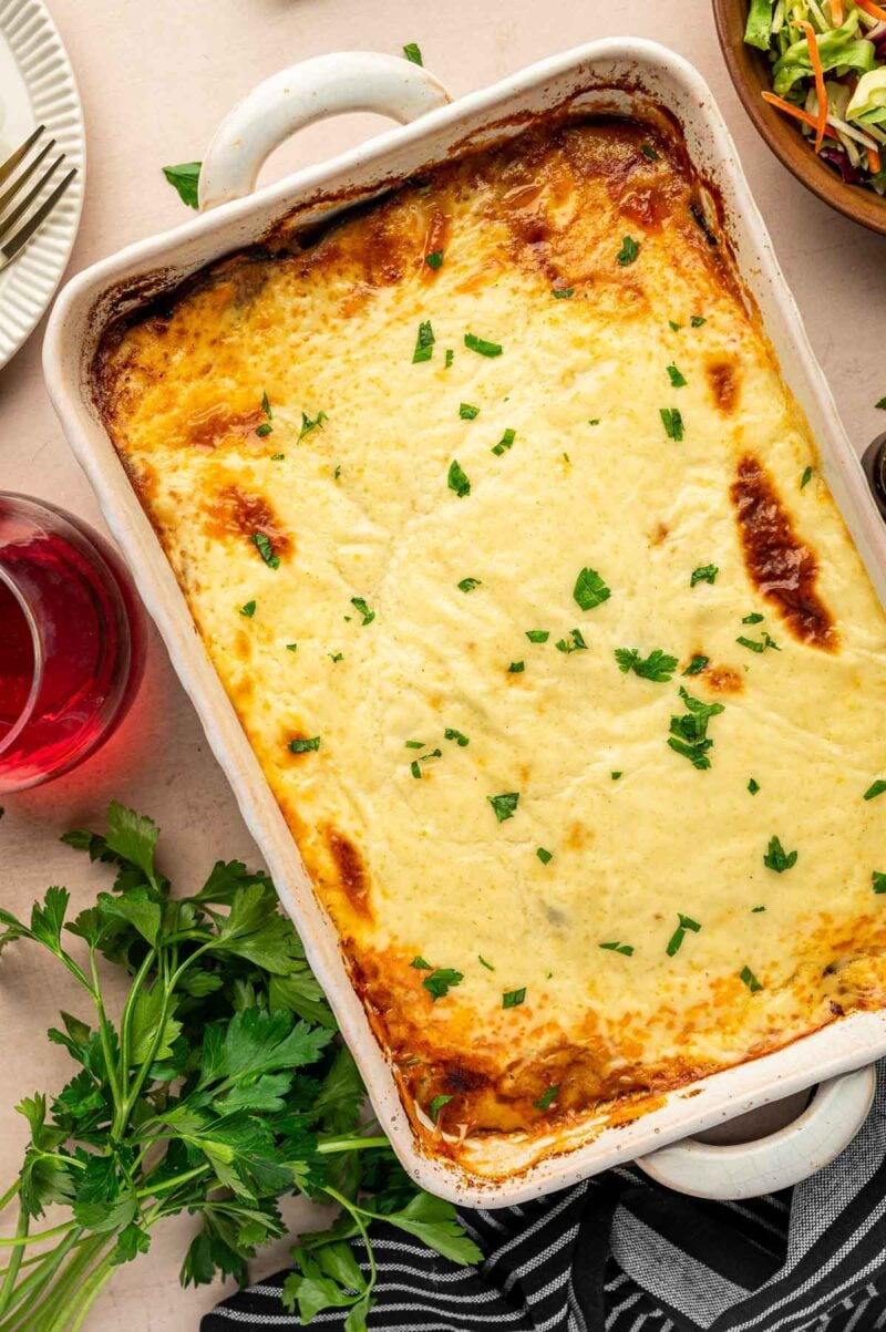 A baked lamb moussaka topped with melted cheese and sprinkled with chopped parsley sits on a table next to fresh herbs, a drink, and a salad.