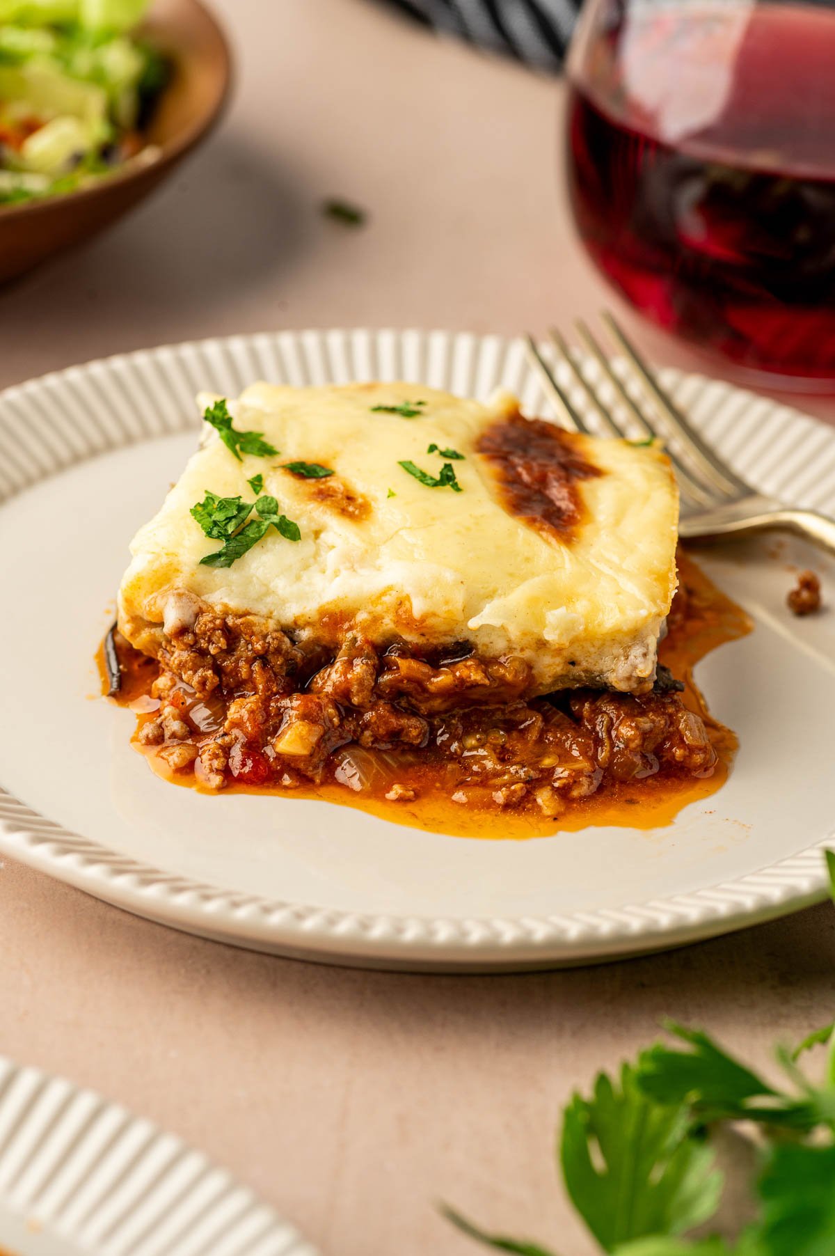 A slice of moussaka with ground lamb, tomato sauce, and creamy béchamel on a white plate, garnished with parsley, with a fork and glass of red wine nearby.