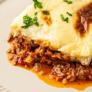 A slice of Moussaka with meat sauce, creamy cheese topping, and garnished with chopped parsley on a white plate.