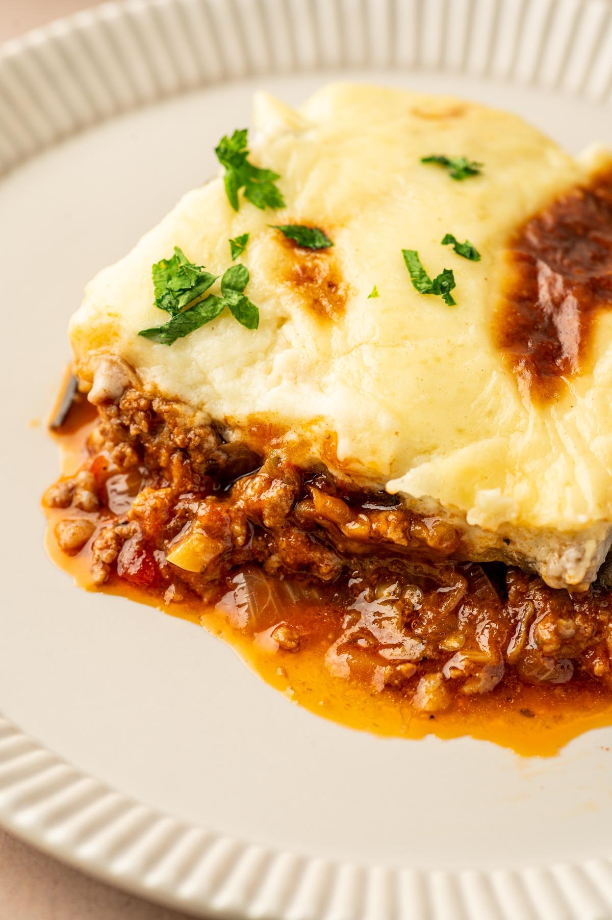 A slice of Moussaka with meat sauce, creamy cheese topping, and garnished with chopped parsley on a white plate.