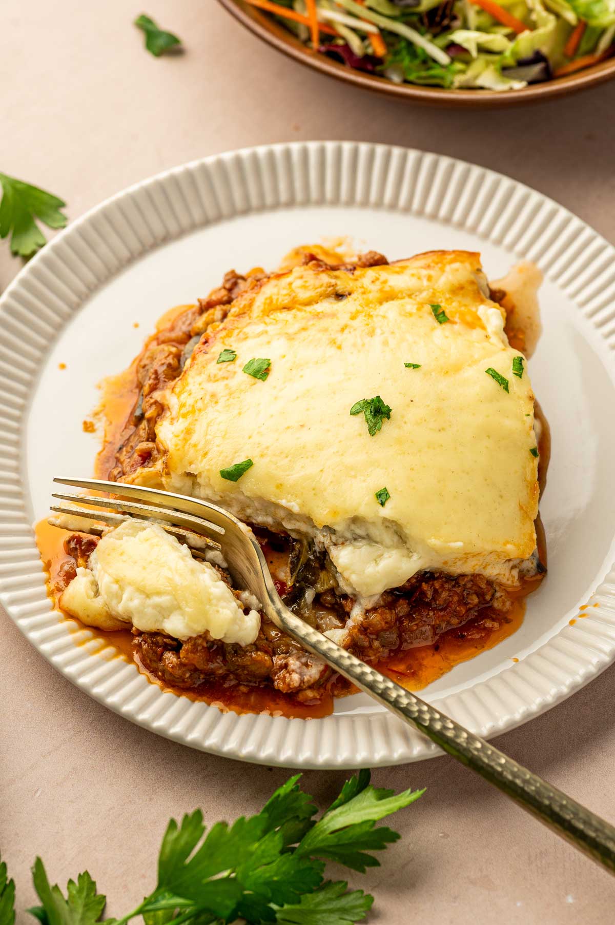 A plate of moussaka with a fork cutting into it, showing layers of meat sauce, pasta, and melted cheese; salad and parsley garnish in the background.
