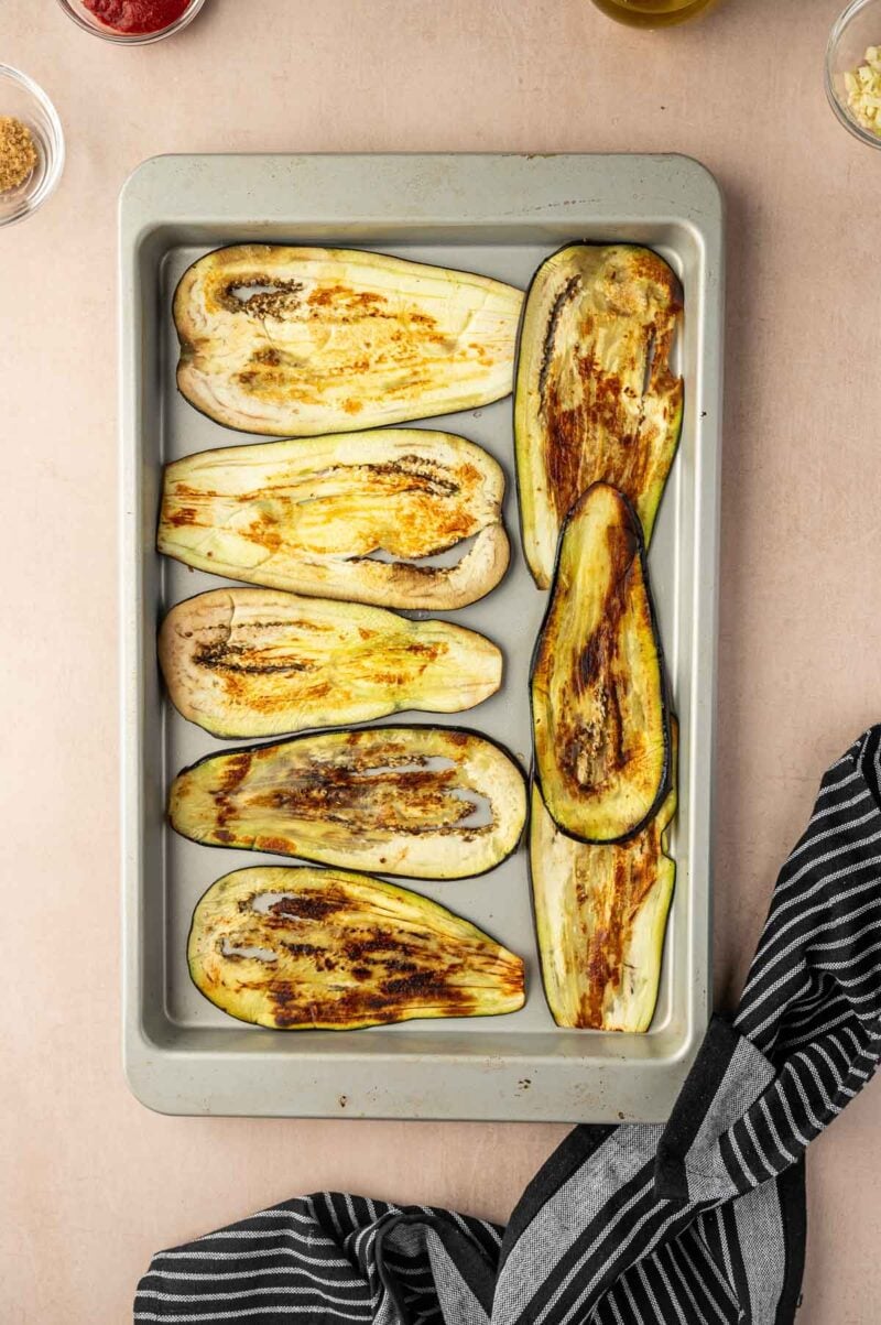 A baking tray with eight roasted eggplant halves, slightly browned, resting on a beige surface with a black and white striped kitchen towel nearby.