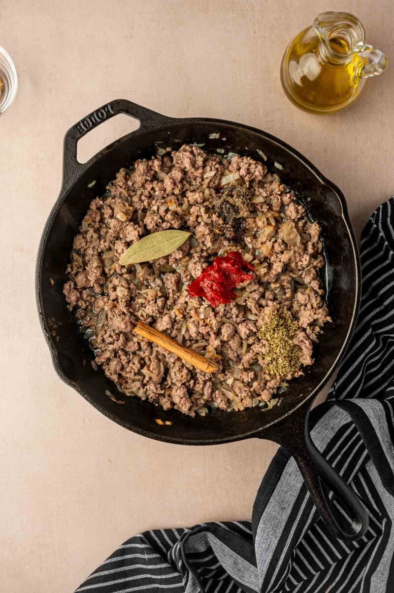 Ground lamb cooking in a cast iron skillet with a bay leaf, tomato paste, cinnamon stick, black pepper, and herbs; a striped towel and a bottle of olive oil are nearby.