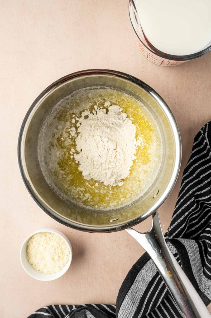 A saucepan with melted butter and flour, a measuring cup of milk, a bowl of grated cheese, and a black-and-white striped towel on a beige surface.