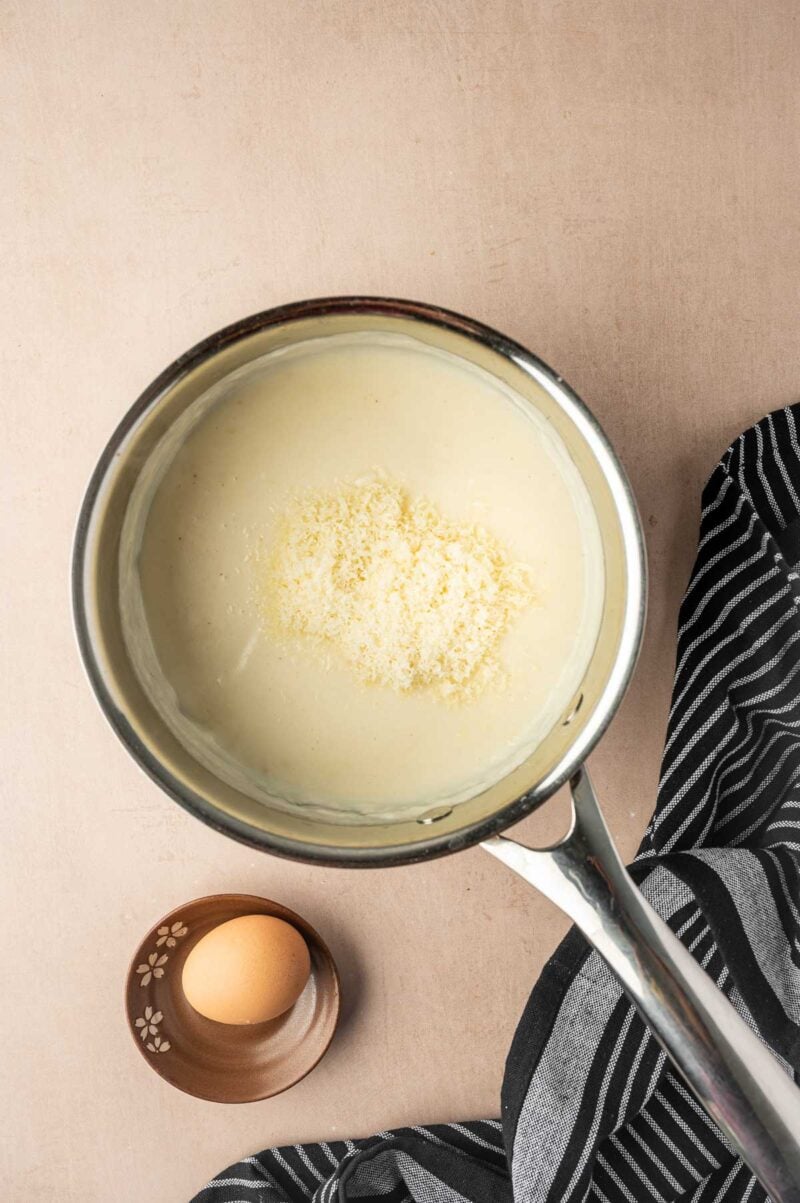 A saucepan filled with creamy white sauce topped with grated cheese sits beside a bowl containing a whole egg. A striped cloth is partially visible next to the pan.