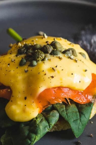 An English muffin topped with spinach, smoked salmon, poached egg, hollandaise sauce, capers, and black pepper on a dark plate with a fork beside it.