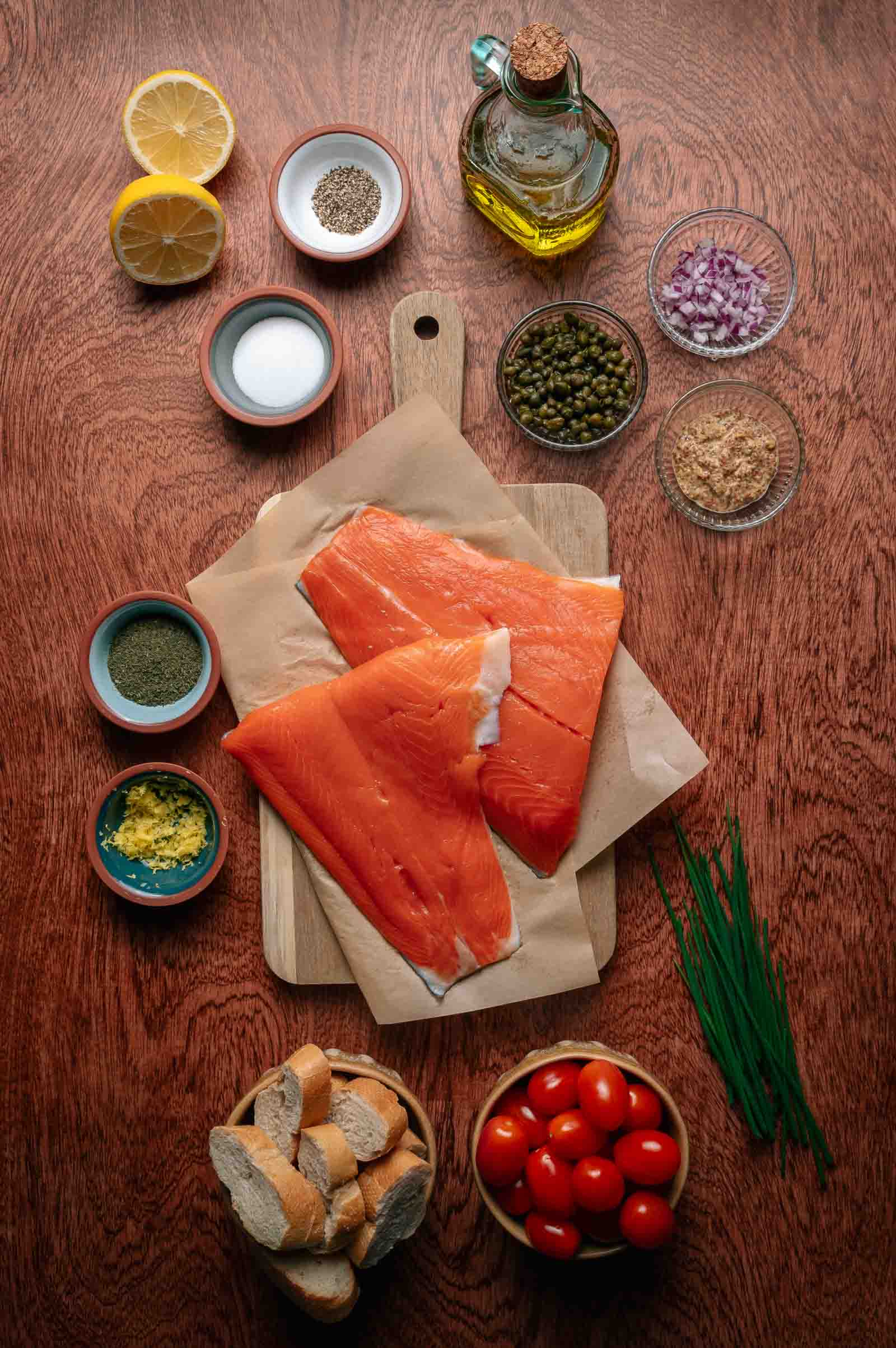 Two raw salmon fillets on parchment paper with bowls of chopped onions, capers, spices, lemon halves, cherry tomatoes, bread slices, oil, and fresh chives arranged on a wooden table.