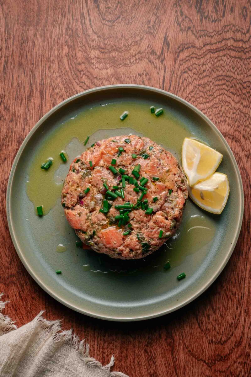 A round serving of salmon tartare garnished with chopped chives, drizzled with oil, and accompanied by two lemon wedges on a green plate.