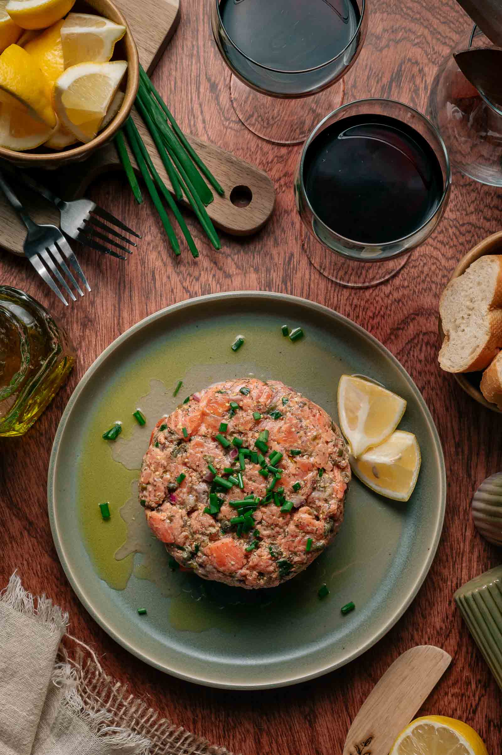 A plate with salmon tartare garnished with chopped chives and lemon wedges, surrounded by wine glasses, bread slices, lemon, and utensils on a wooden table.