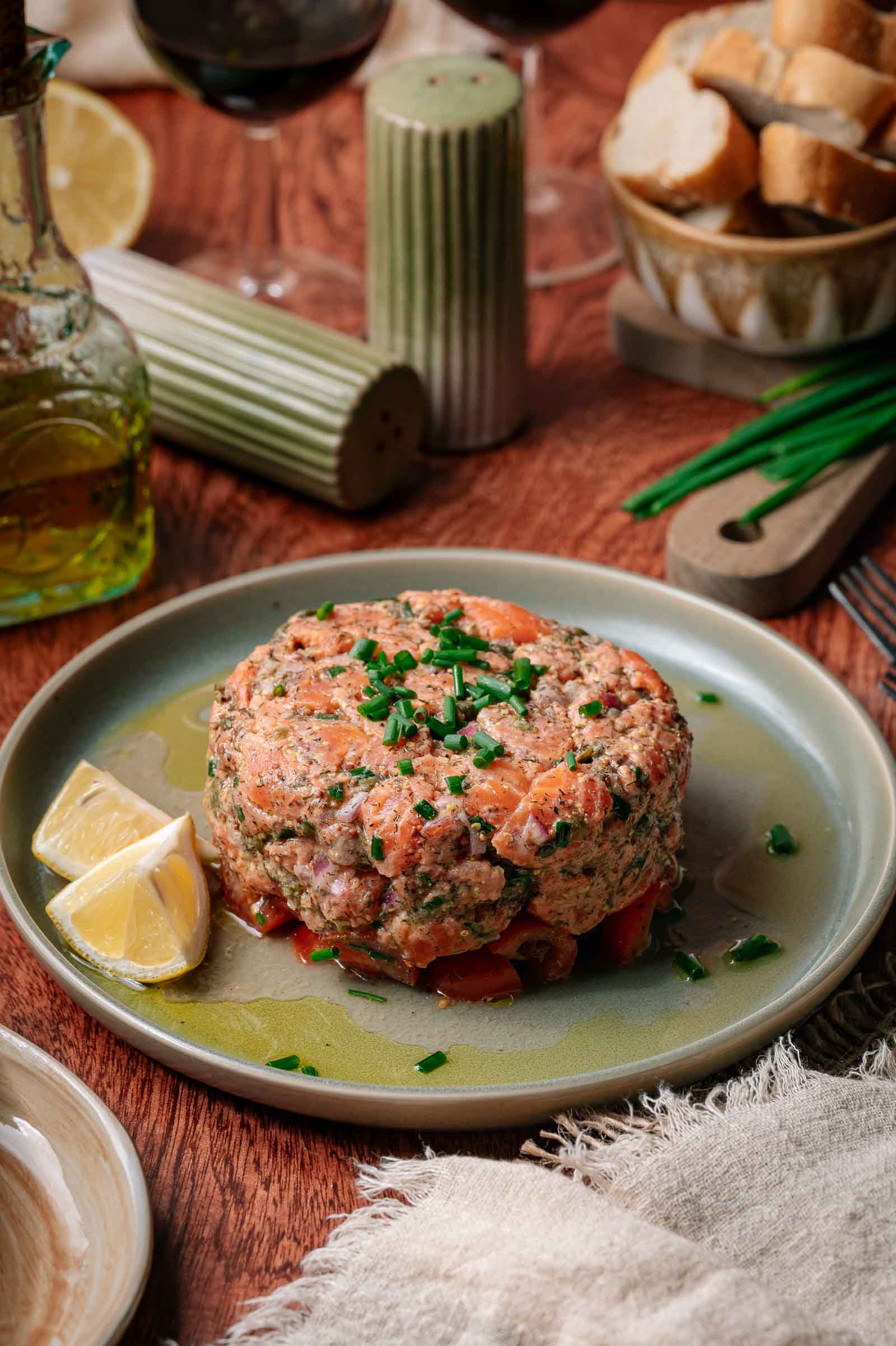 A plate with salmon tartare garnished with chopped chives and lemon wedges, surrounded by bread, olive oil, and tableware on a wooden table.