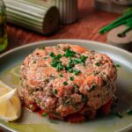 A round serving of salmon tartare garnished with chopped chives sits on a plate with lemon wedges, on a wooden table with condiments in the background.