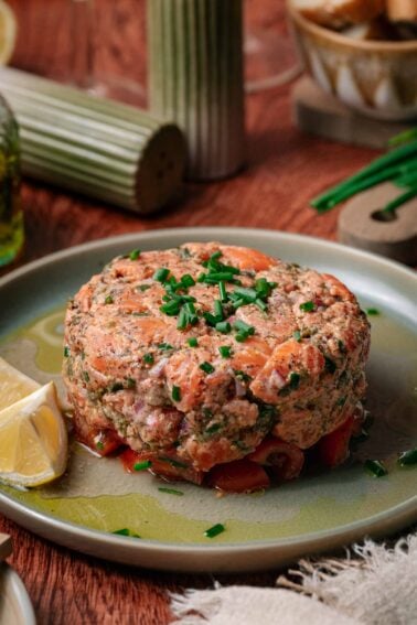 A round serving of salmon tartare garnished with chopped chives sits on a plate with lemon wedges, on a wooden table with condiments in the background.