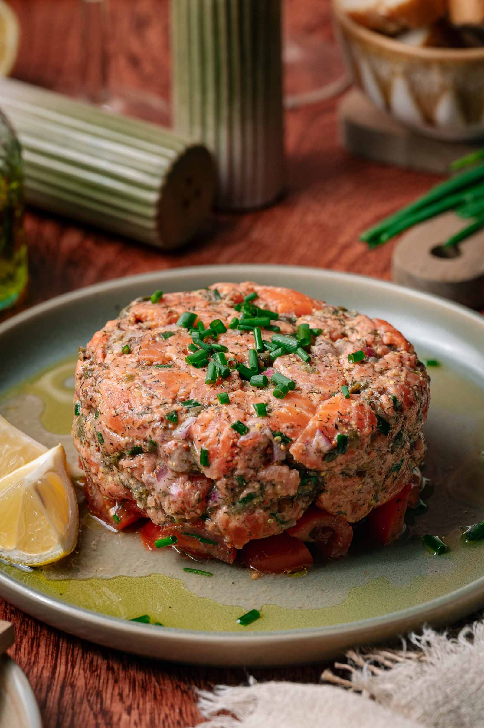 A round serving of salmon tartare garnished with chopped chives sits on a plate with lemon wedges, on a wooden table with condiments in the background.
