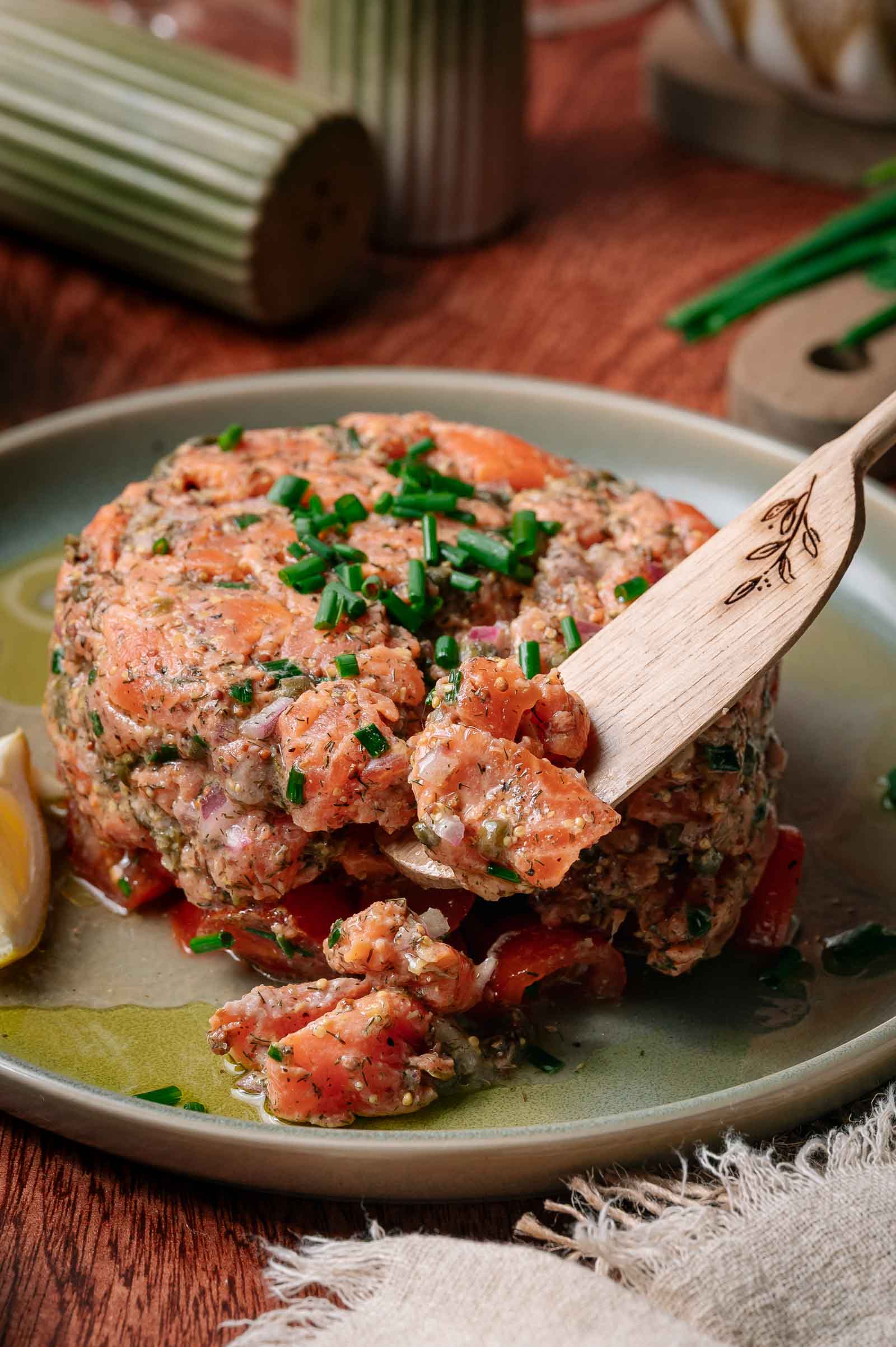 A close-up of a serving of salmon tartare with herbs and diced vegetables, garnished with chopped chives on a green plate, with a wooden fork and a lemon wedge beside it.