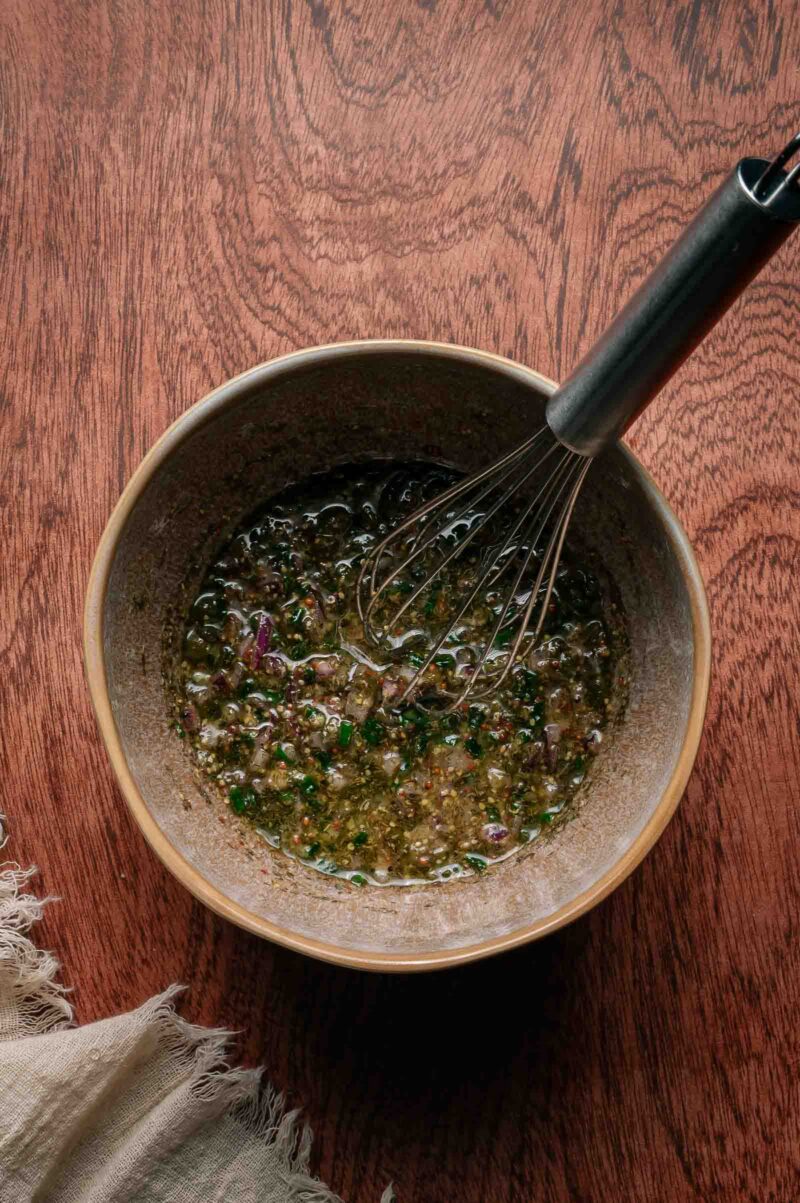 A small bowl of mustard and oil with finely chopped herbs and a metal whisk, set on a wooden surface.
