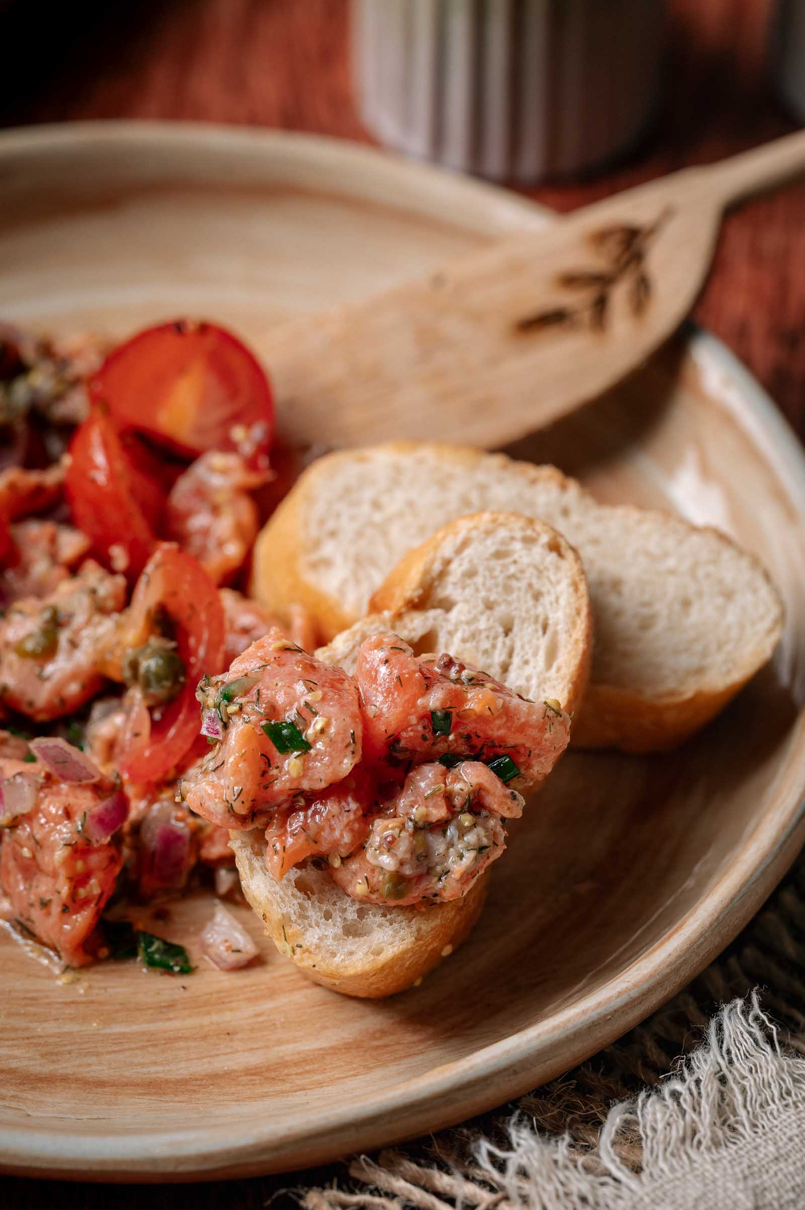 A plate with sliced baguette and a salmon tartare topping containing diced tomatoes, herbs, and onions, with a wooden spoon in the background.