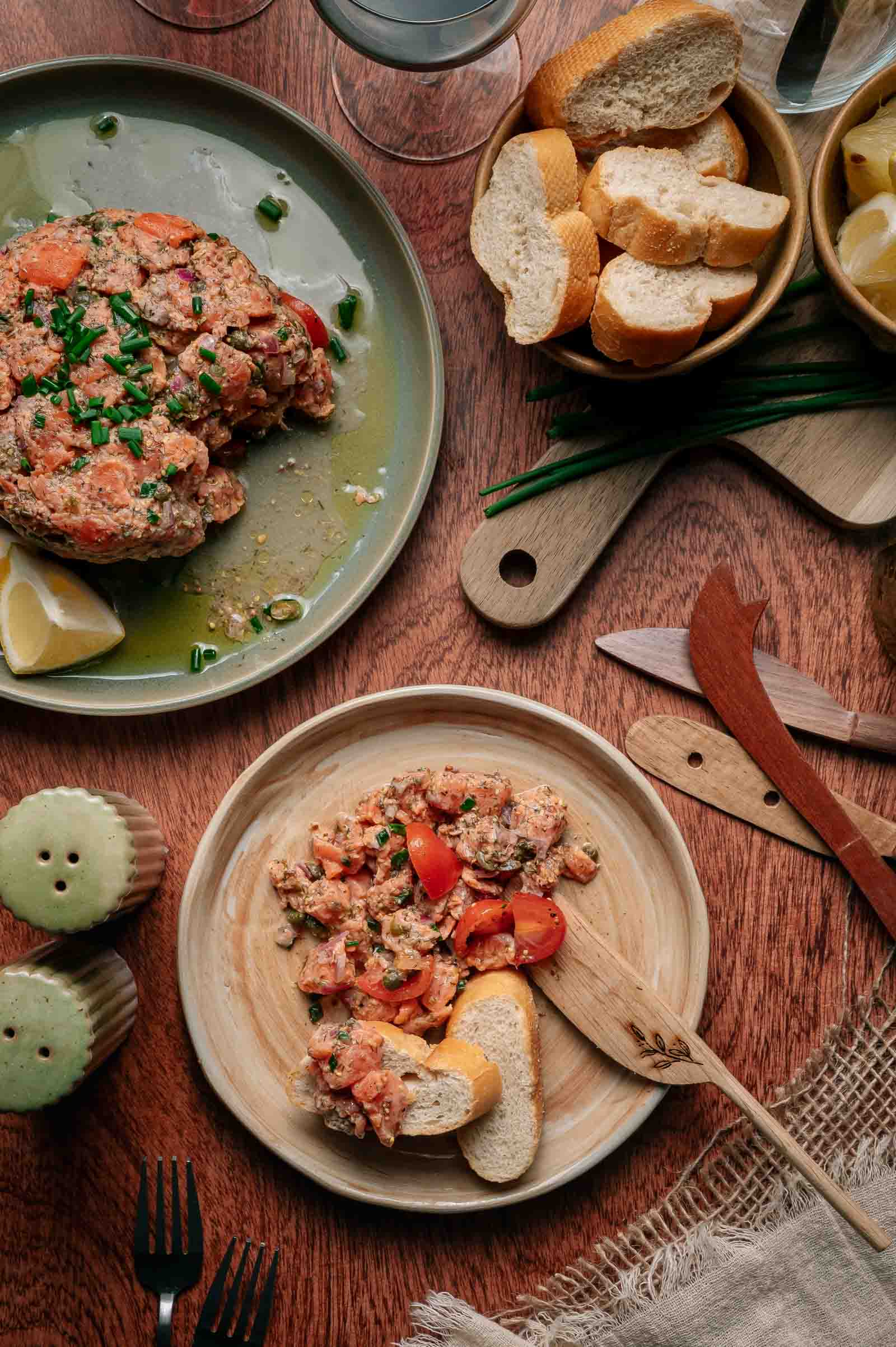 Two plates of salmon tartare with chopped tomatoes and herbs, served with lemon wedges and sliced baguette on a wooden table set with utensils and garnishes.