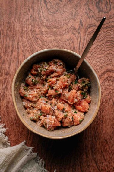 A brown ceramic bowl filled with a mixture of diced raw salmon, herbs, and seasonings, with a spoon resting inside, placed on a wooden surface.