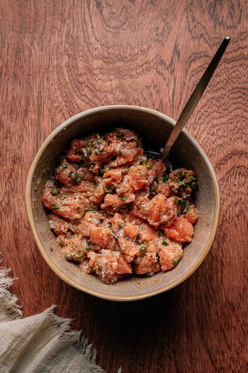 A brown ceramic bowl filled with a mixture of diced raw salmon, herbs, and seasonings, with a spoon resting inside, placed on a wooden surface.