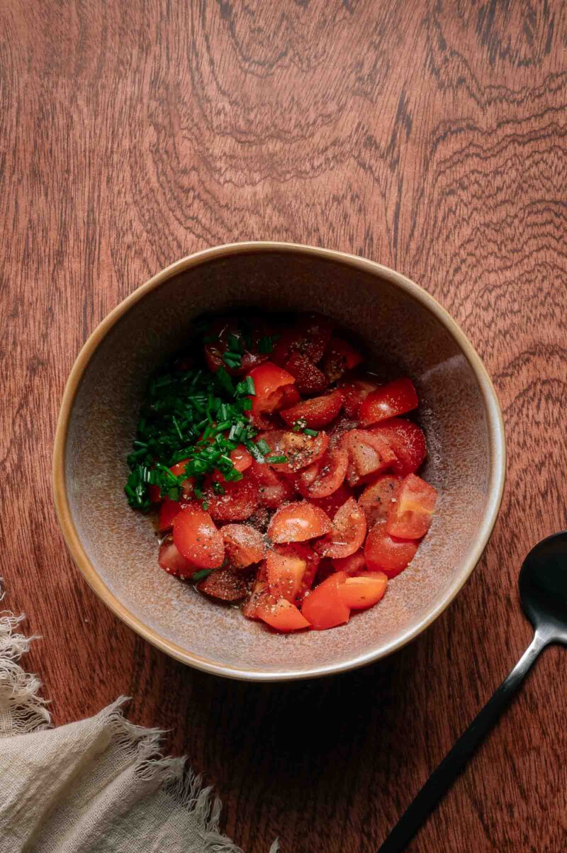 A bowl of chopped tomatoes and herbs sprinkled with black pepper sits on a wooden table next to a spoon and a fringed cloth.