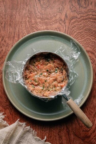 A metal pot lined with plastic wrap containing a mixture of salmon tartare and herbs sits on a green plate on a wooden surface.