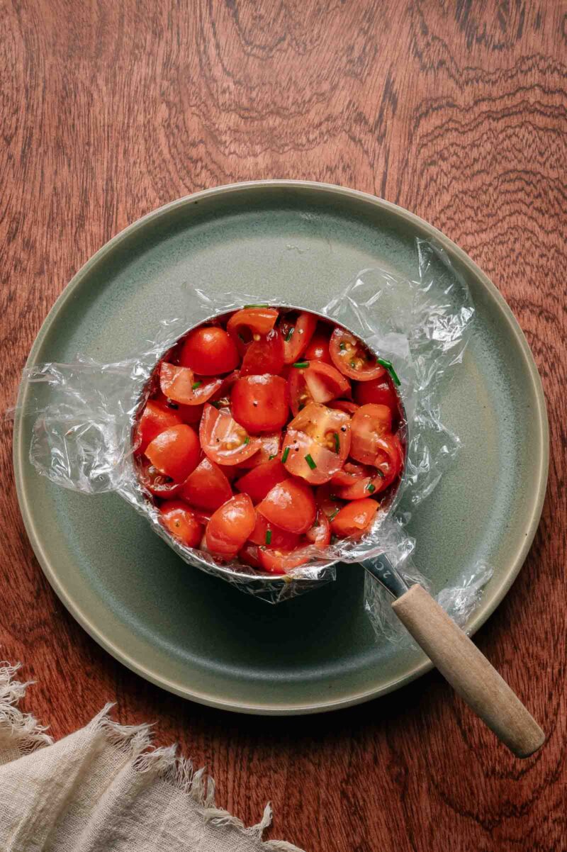 A green plate holds a saucepan lined with plastic wrap, filled with chopped cherry tomatoes and herbs on a wooden table.