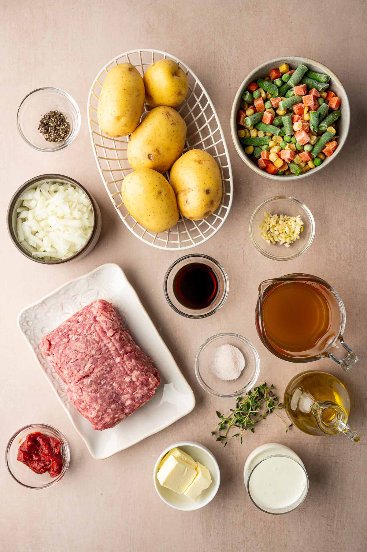 Top-down view of uncooked ingredients for a shepherd's pie, including potatoes, ground meat, mixed vegetables, broth, onions, garlic, butter, cream, tomato paste, seasonings, and herbs.