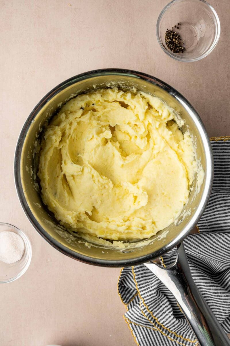 A pot of creamy mashed potatoes sits on a counter next to a striped kitchen towel, with small bowls of salt and pepper nearby.