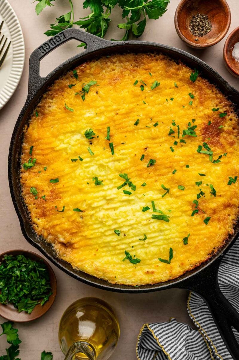 A baked shepherd’s pie in a cast iron skillet, topped with golden mashed potatoes with crispy ridges and garnished with chopped parsley.