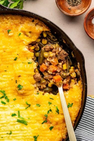 A skillet of shepherd's pie with a spoon scooping out a portion, showing ground lamb, peas, corn, and carrots under a mashed potato topping.